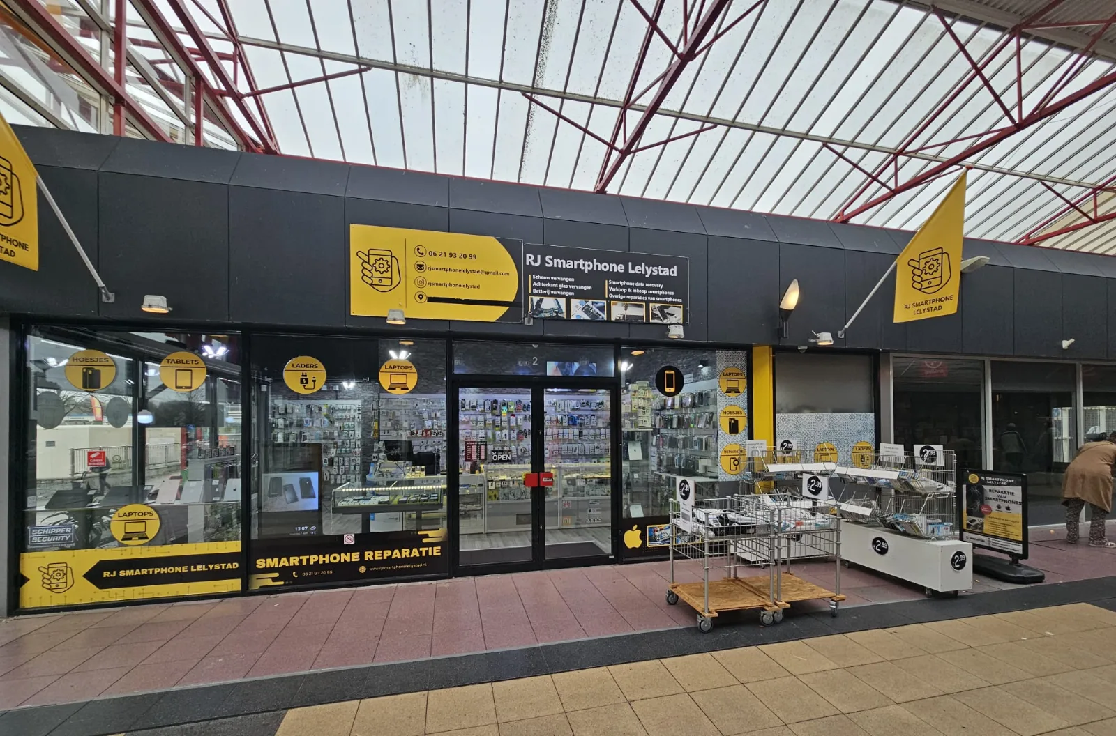De voorgevel van RJ Smartphone Lelystad aan de Weversstraat 2: een donkere pui met geel logo, reclameborden met reparaties en de etalage met toestellen